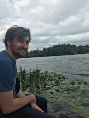 Connor sitting next to the water, smiling with a scenic background of a lake and lush greenery.